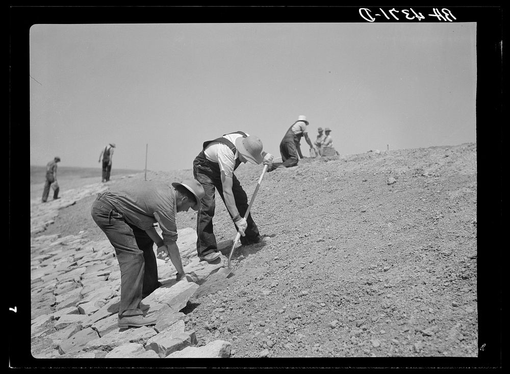 Laying stone on the face of a stock water dam. Pine Ridge land use project. Sioux County, Nebraska. Sourced from the Library…