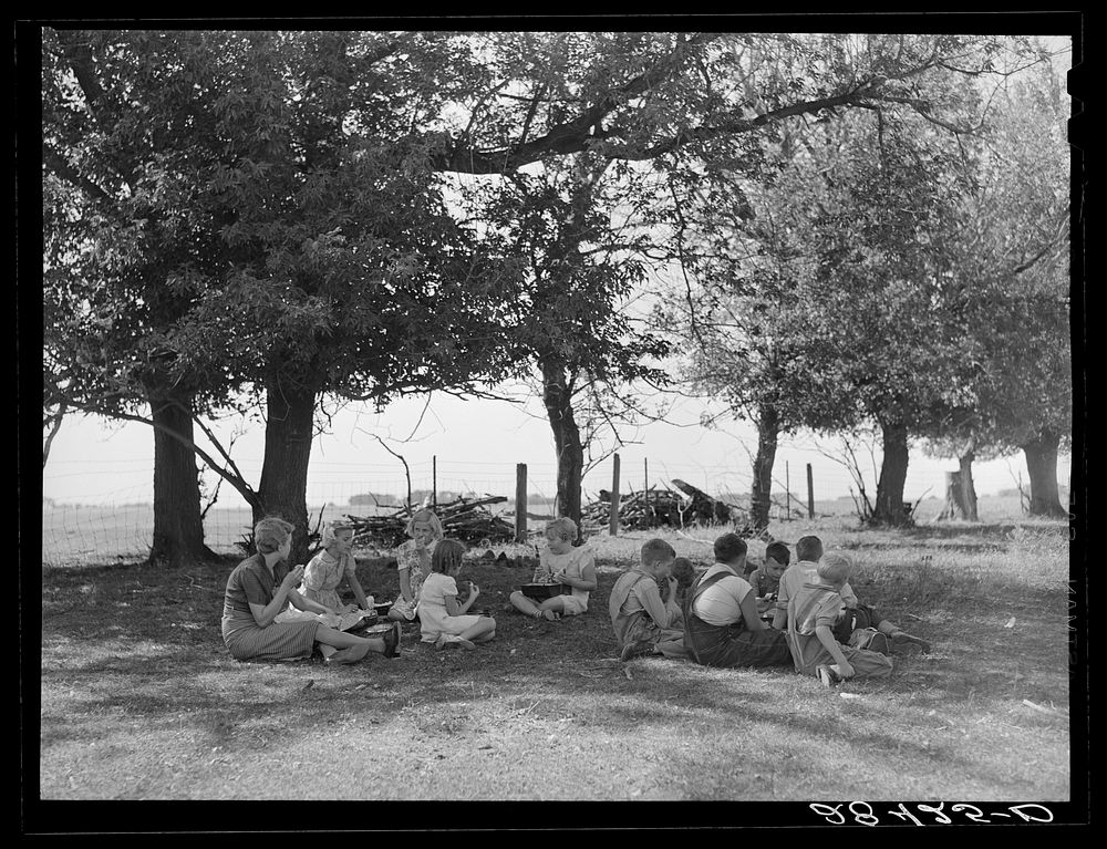 Teacher eats lunch with children in shade of trees on school grounds. Grundy County, Iowa. Sourced from the Library of…