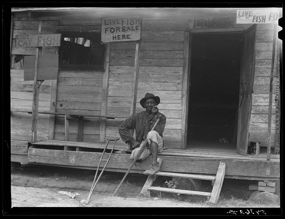 Natchitoches, Louisiana. A  sitting on the porch of a store where live fish are for sale. Sourced from the Library of…