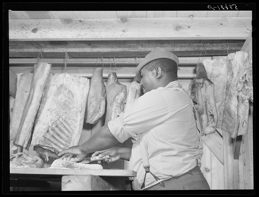 Lewis Wiggins cutting off a slice of homecured ham in his smokehouse. La Delta Project, Thomastown, Louisiana. Sourced from…
