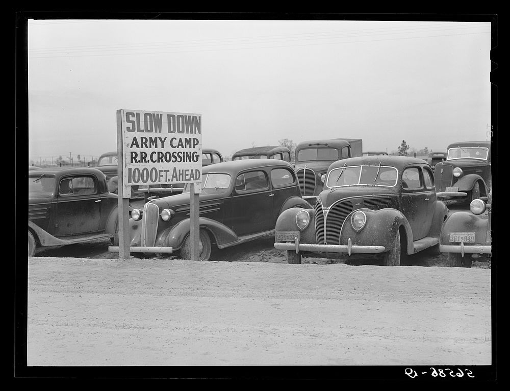 Cars of construction workers parked along highway by entrance to Camp Claiborne. Near Alexandria, Louisiana. Sourced from…