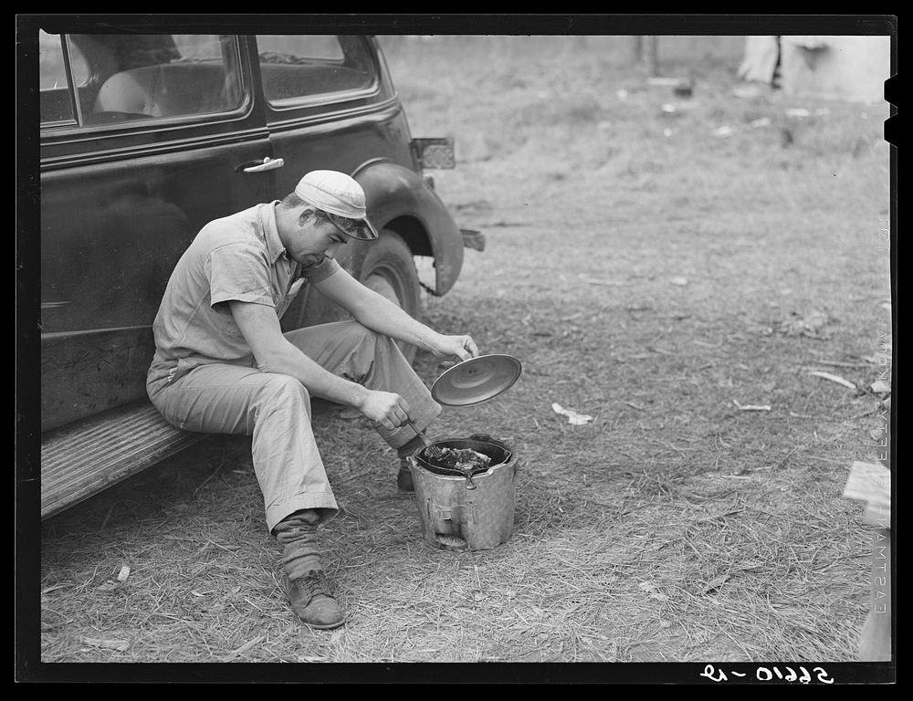 John H. Poole, Jr. cooking while sitting on side of car. He is a carpenter on construction job at Camp Livingston. He comes…