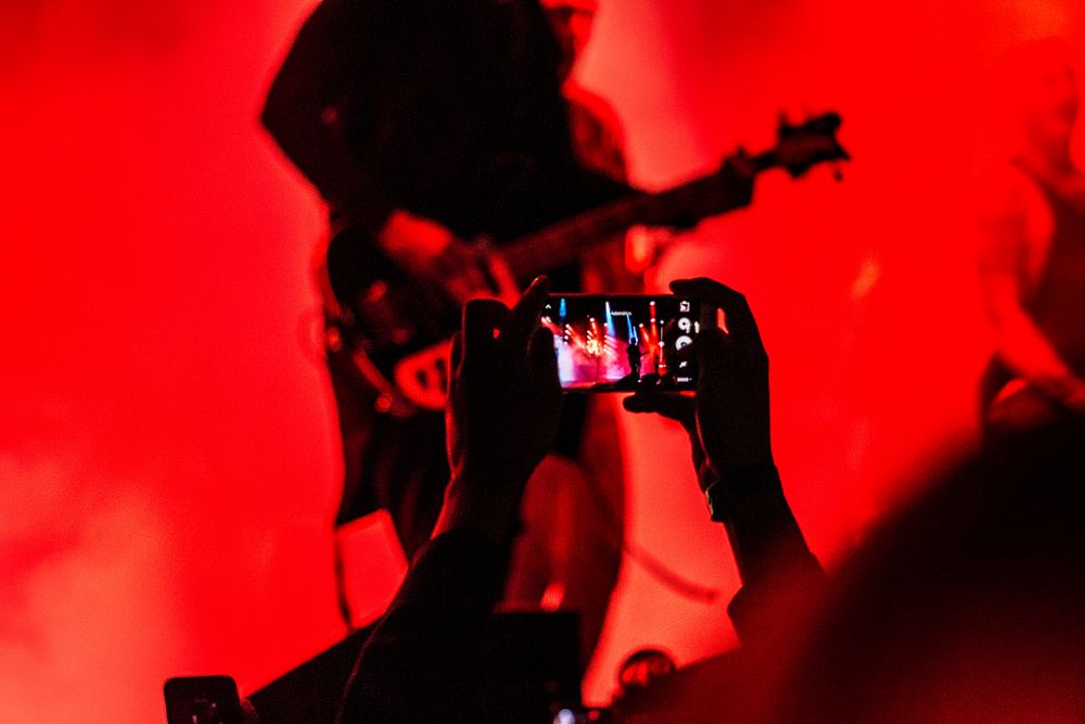 A guitarist performs on stage in red light while an audience member records a concert on their phone. Original public domain…