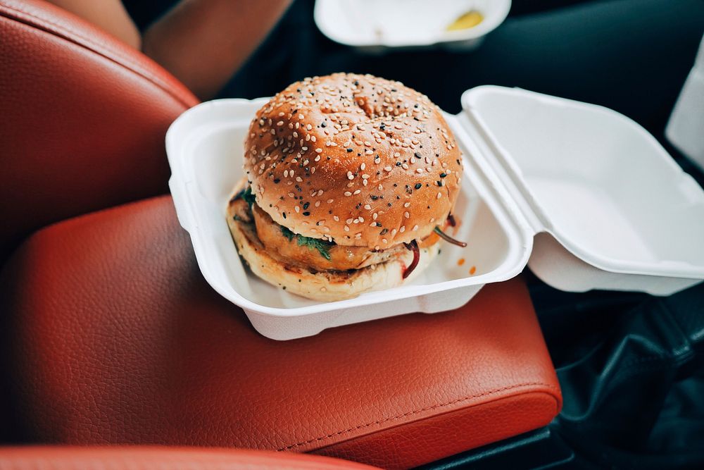 Burger with a sesame bun in a styrofoam container on the center console in a car. Original public domain image from…