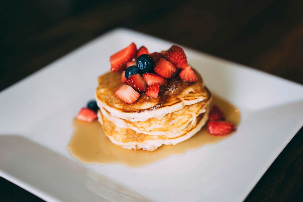 A stack of golden brown pancakes covered in syrup and fresh strawberries and blueberries on a white plate. Original public…