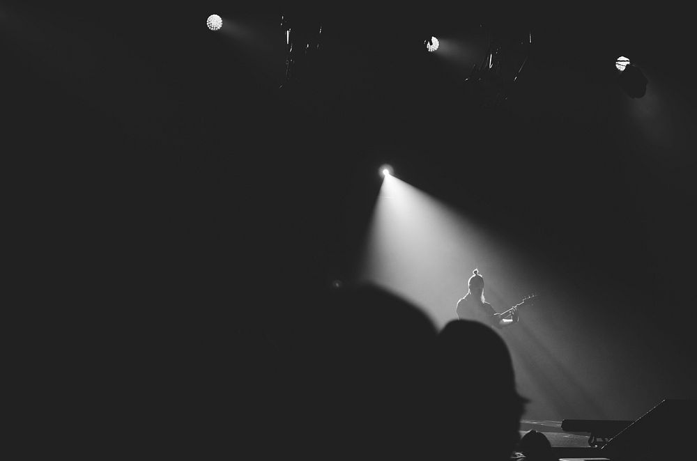A guitar player illuminated by white floodlights in the middle of a dark stage. Original public domain image from Wikimedia…