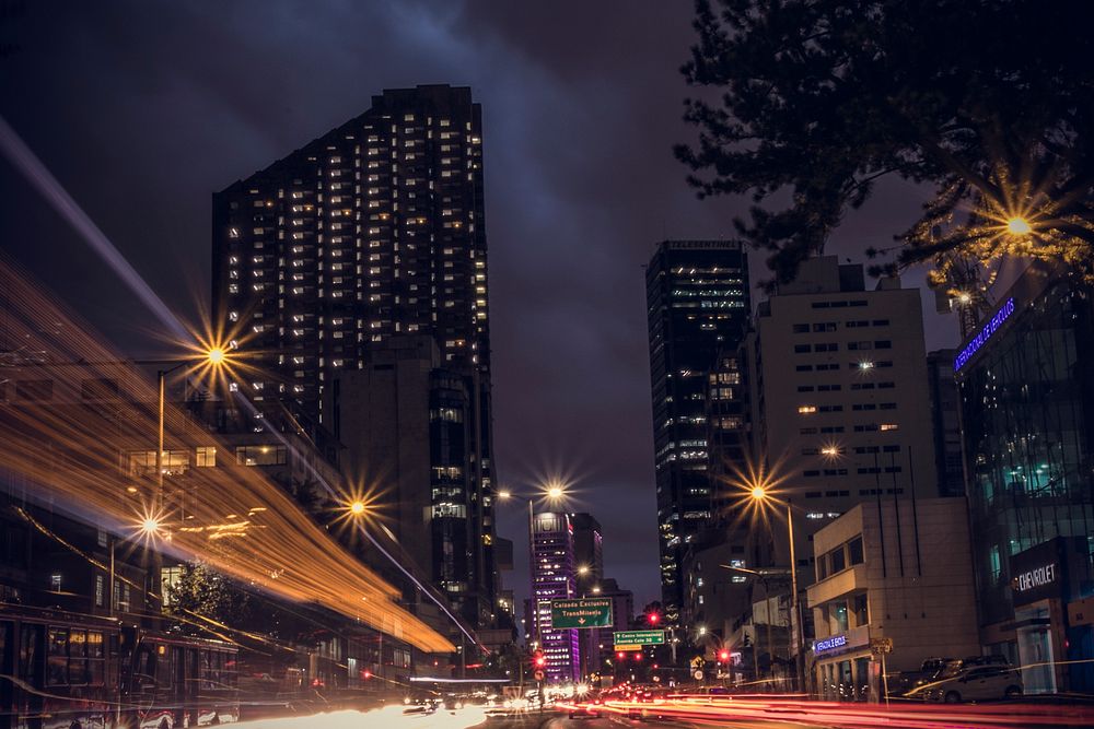 A dark city background featuring large buildings and street light trails in Bogotá. Original public domain image from…