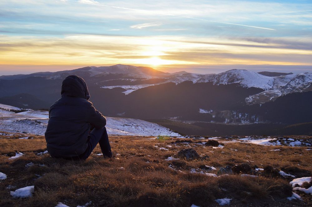 A person sitting atop a mountain in Sinaia, watching the sunset above snowy mountain range. Original public domain image…