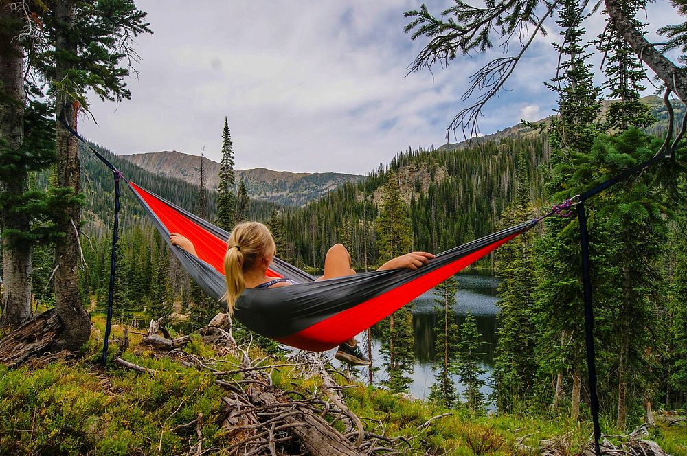 A girl with blonde hair sits in a red and gray hammock overlooking mountains, trees, and water.. Original public domain…