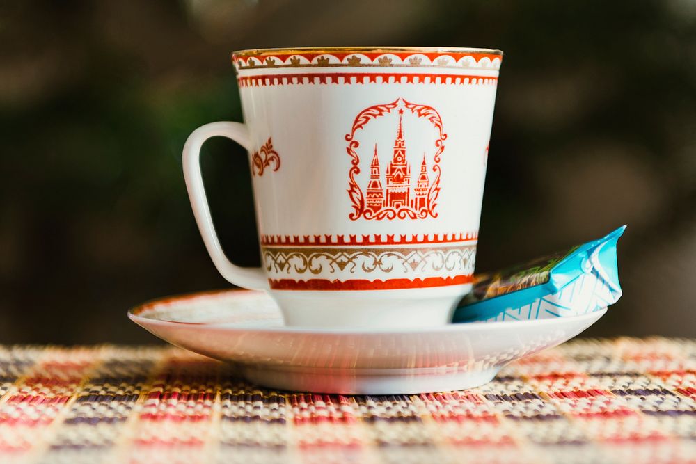A ceramic cup with a castle on it sitting on a saucer with a biscuit in a wrapper. Original public domain image from…