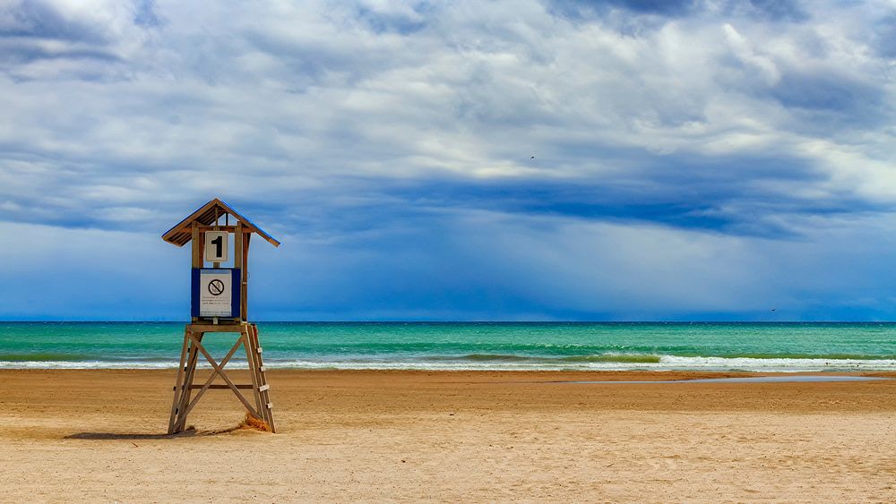 A lifeguard stand on a deserted beach along a green ocean under a blue sky. Original public domain image from Wikimedia…