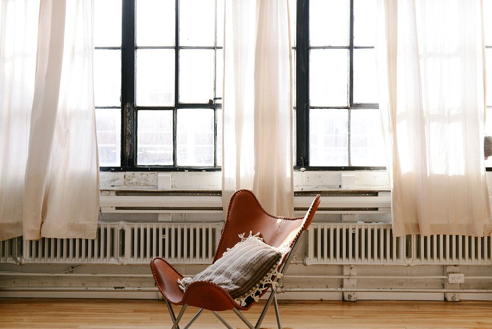 A brown leather chair with a throw pillow on it, in a big, light and airy room with white curtains in Breather Montreal.…