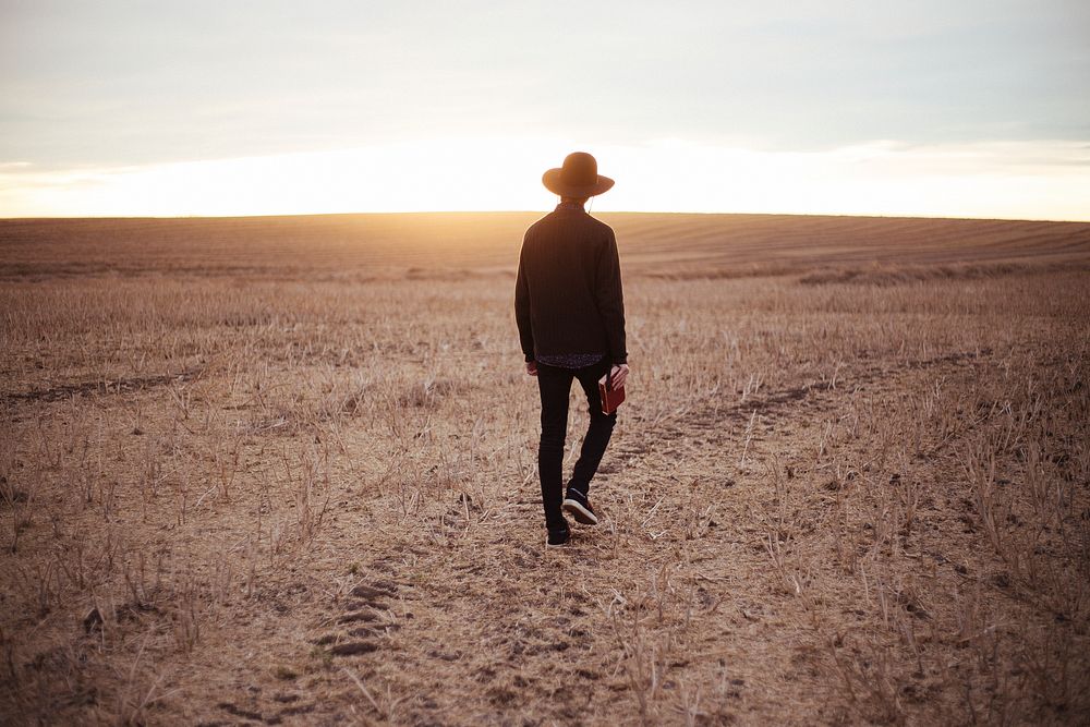 A guy wearing a hat and holding a book, walking through a field during golden hour. Original public domain image from…