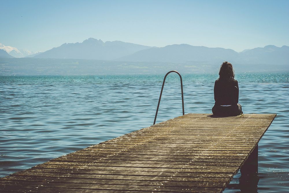 A girl in Morges sits at the end of a dock and stares at the mountains. Original public domain image from Wikimedia Commons