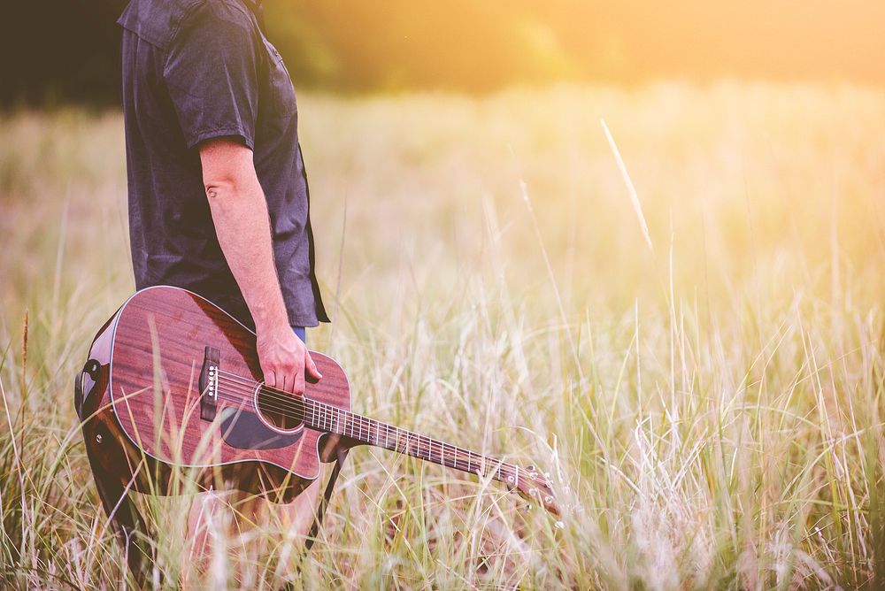 A man holding brown acoustic guitar standing on dried grass field. Original public domain image from Wikimedia Commons