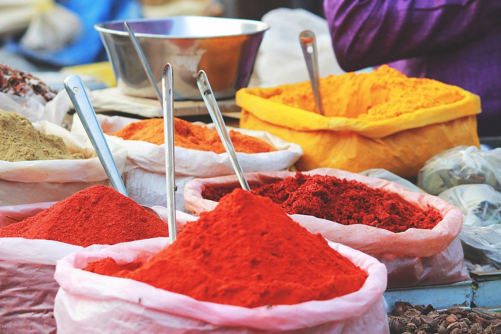 Red, tan, yellow, and orange spices in plastic bags with scooping spoons in a marketplace. Original public domain image from…