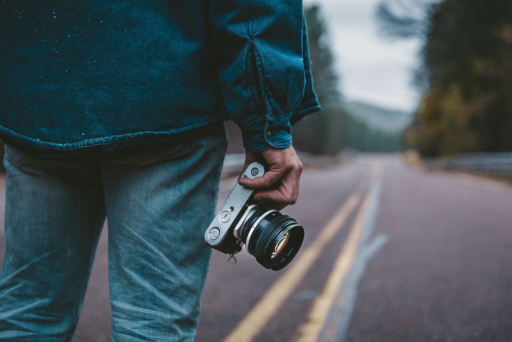 A man in denim clothing stands on an empty paved road holding a camera. Original public domain image from Wikimedia Commons