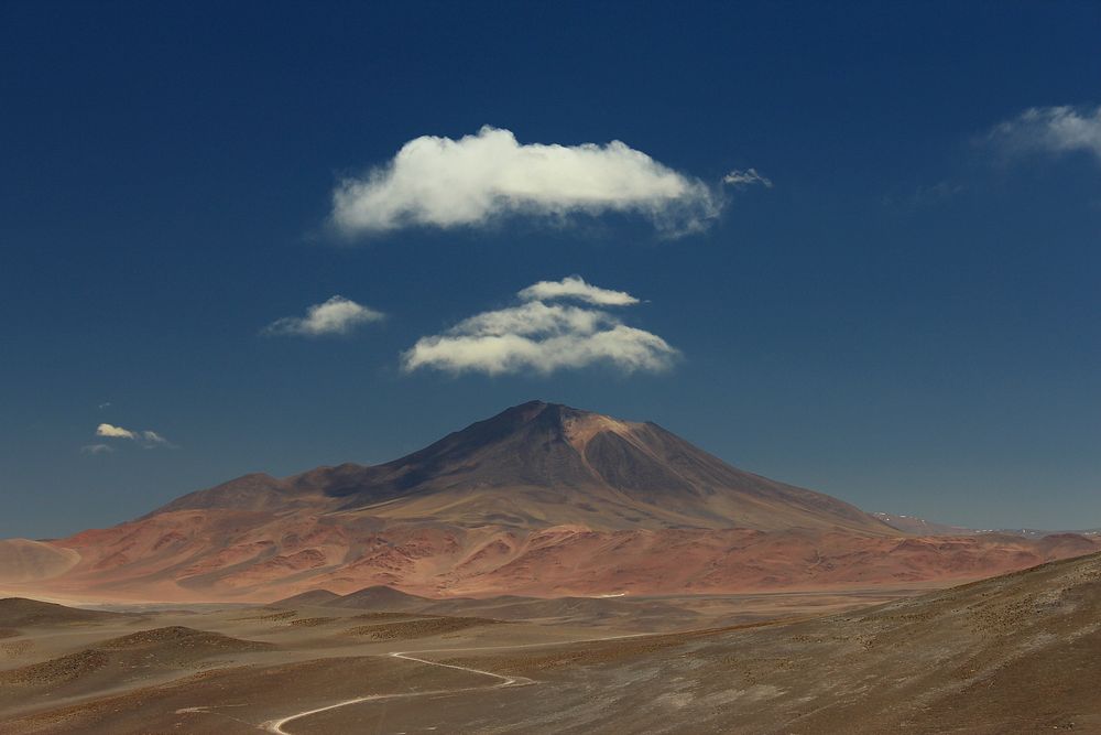 Fluffy clouds over a tall mountain with red rock slopes in Salar de Incahuasi. Original public domain image from Wikimedia…