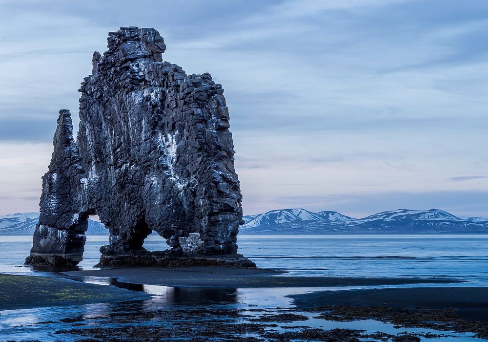 Tall rock formation above the ocean with snow covered mountains in the background at Hvítserkur. Original public domain…