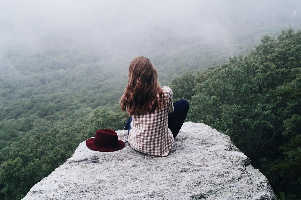 A lone woman sitting on a rocky ledge overlooking a forest with a red hat on her side. Original public domain image from…