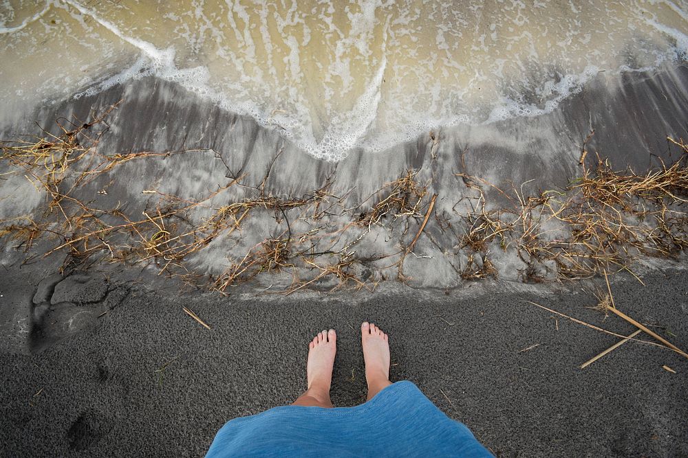 An overhead shot of a person's feet on gray sand on a wild beach. Original public domain image from Wikimedia Commons