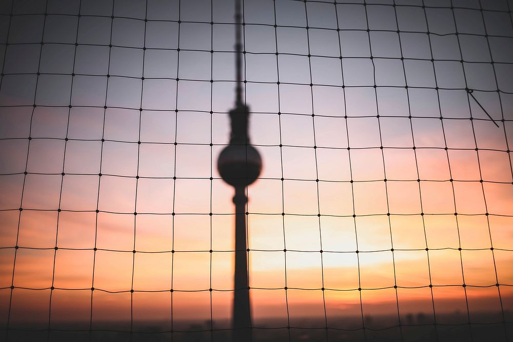 A silhouette of the Fernsehturm tower through netting, at sunset in Berlin. Original public domain image from Wikimedia…