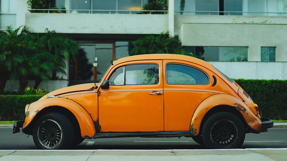 Rusted old vintage orange Volkswagen beetle parked in front of apartment complex. Original public domain image from…