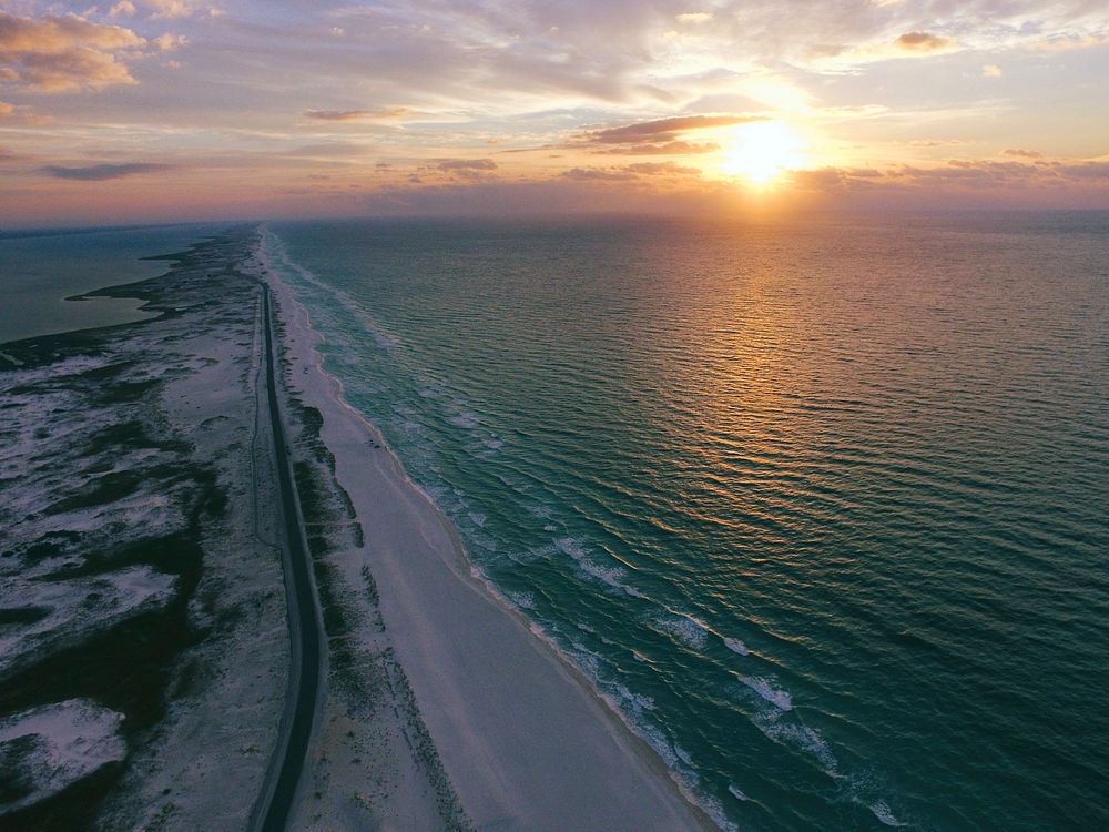 Drone view of an empty coastal road beside the beach, sun setting over rippling water. Original public domain image from…