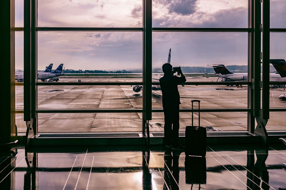 A silhouette of a man taking photos at the floor-to-ceiling window in an airport. Original public domain image from…