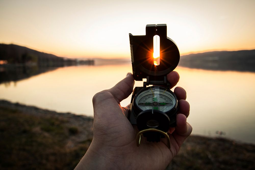 A hand holding up a lensatic compass and pointing the lens at the setting sun. Original public domain image from Wikimedia…