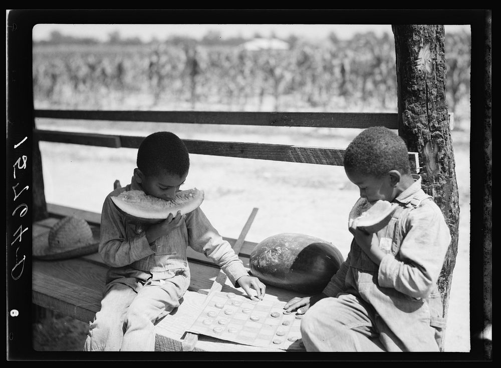  boys eating watermelon and playing checkers with bottle caps. Florence County, South Carolina. Sourced from the Library of…