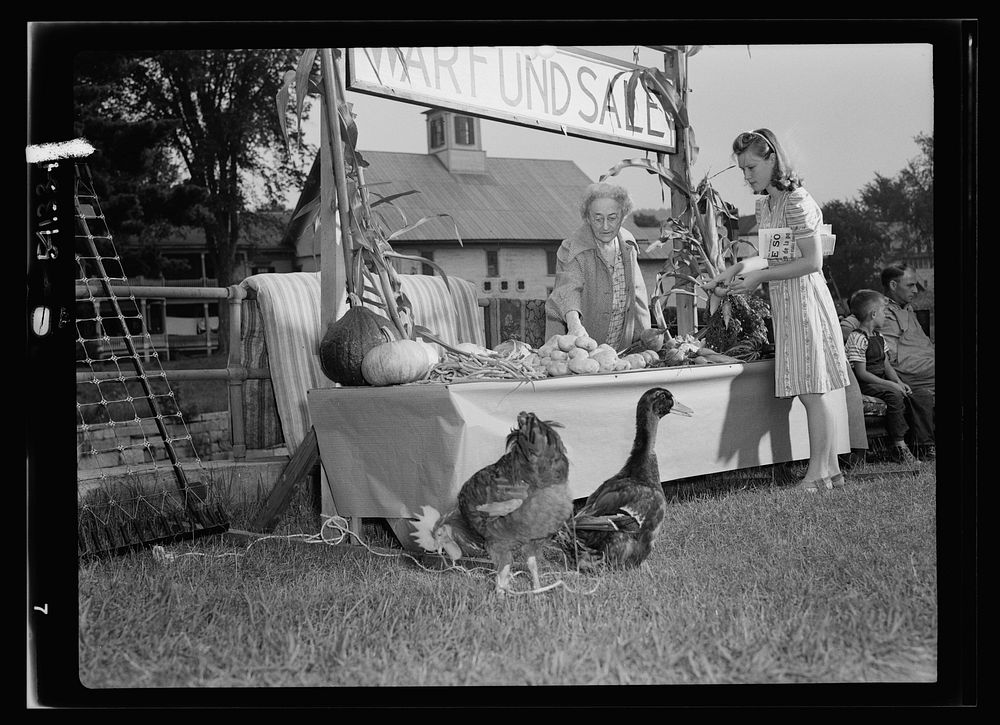 Mrs. Alice White at the Victory Store vegetable counter selling donated farm produce to Miss Lorraine Lavertu. Sourced from…