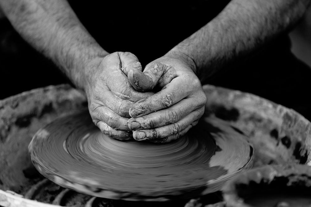 Black and white photo of potter molding clay with his hands on a spinning pottery wheel. Original public domain image from…