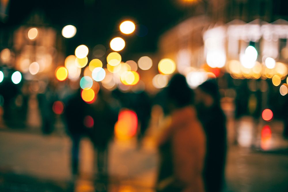 An out-of-focus look at people walking outside in a town at night with with blurs of dotted light all around. Original…