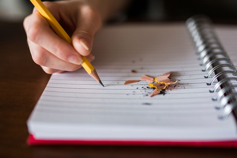 A person writing with a pencil in a notebook with pencil shavings on it. Original public domain image from Wikimedia Commons