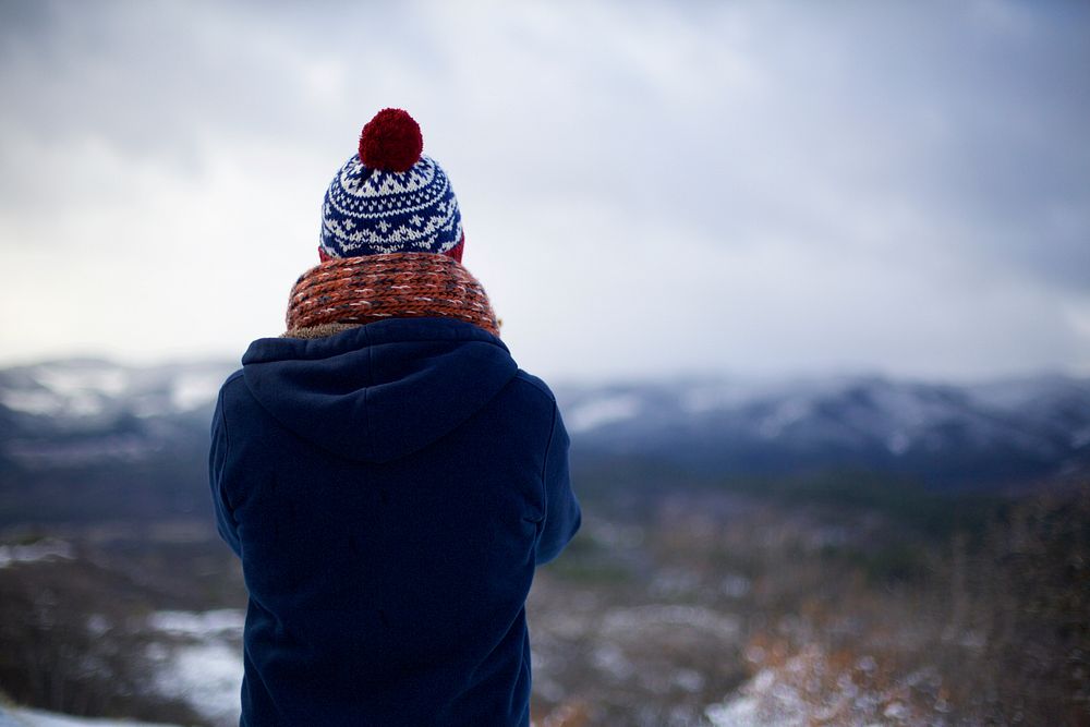 The back of a person with crossed arms wearing a knit cap, scarf, and blue coat is visible as he or she stares out into…
