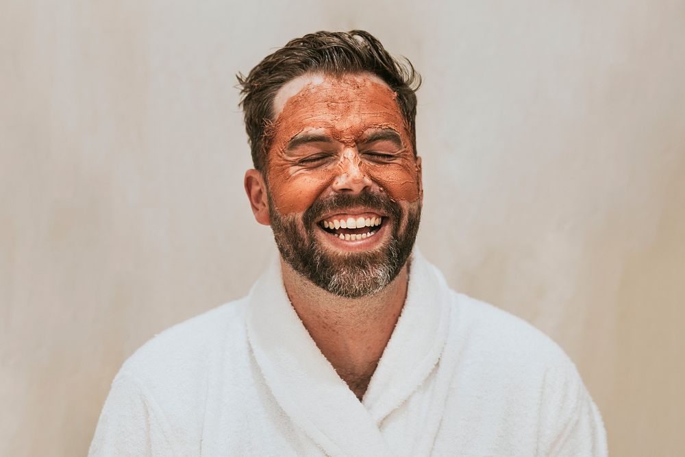 Laughing man in robe with facial mask isolated on background