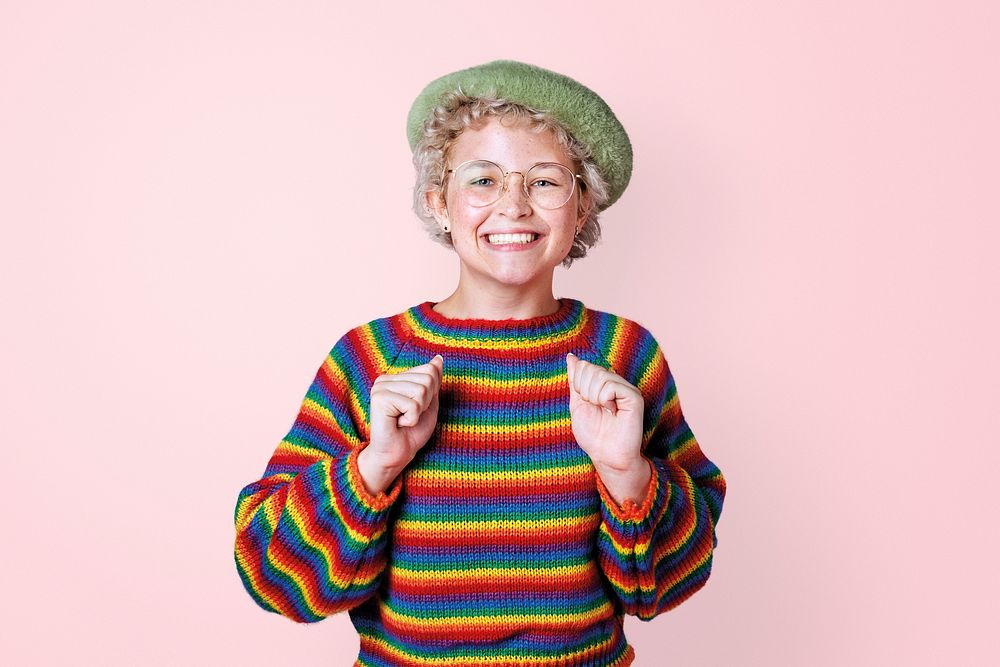 Happy lesbian woman in a rainbow sweater standing by a pink wall 