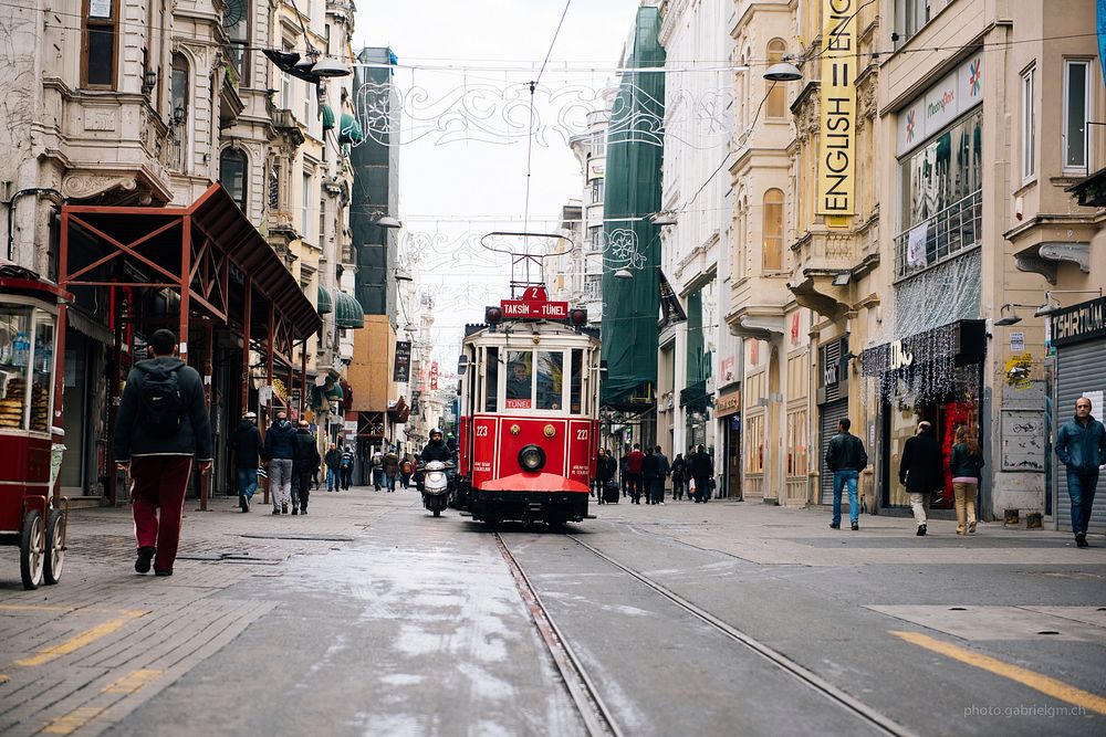 Red cable car in road with people and buildings on either side. Original public domain image from Wikimedia Commons
