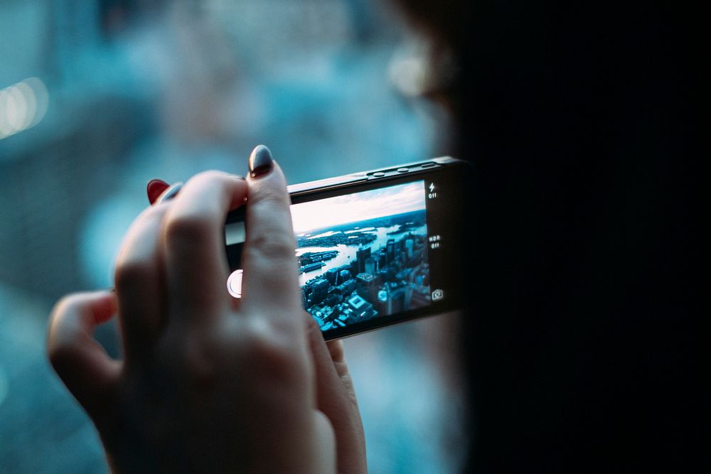Macro of a woman with dark red nails taking a photo of the city on her iPhone. Original public domain image from Wikimedia…