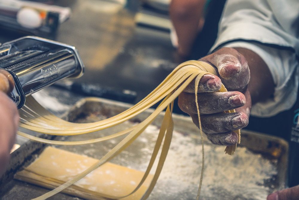 A chef holding long strands of pasta coming out of a machine. Original public domain image from Wikimedia Commons