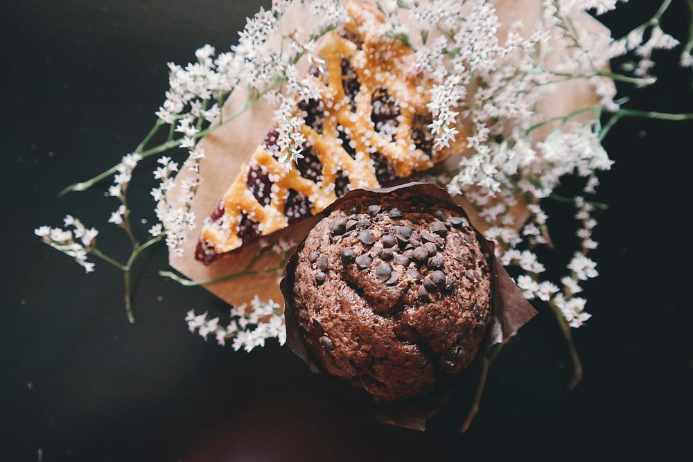 An overhead shot of a chocolate chip muffin next to a piece of pie decorated with elderflowers. Original public domain image…