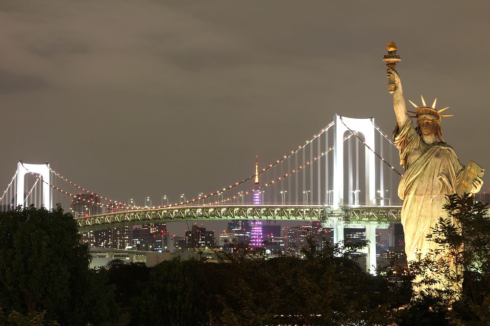 The Statue of Liberty stands in front of a lit up bridge at night in New York City. Original public domain image from…
