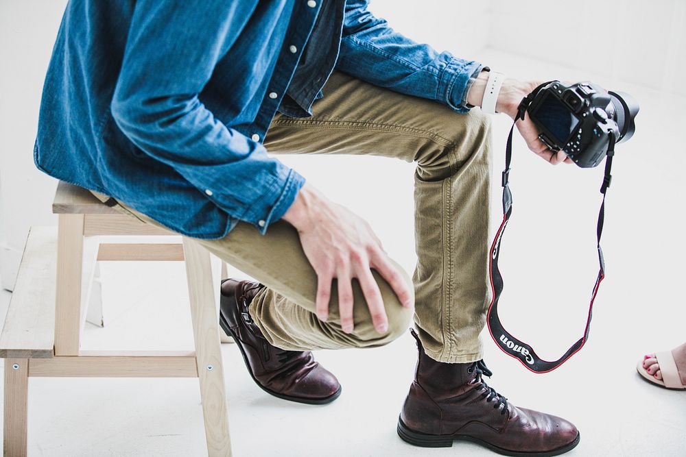 A man wearing a denim shirt and khakis taking a photo with a Canon camera of a woman with sandals and red toenails. Original…