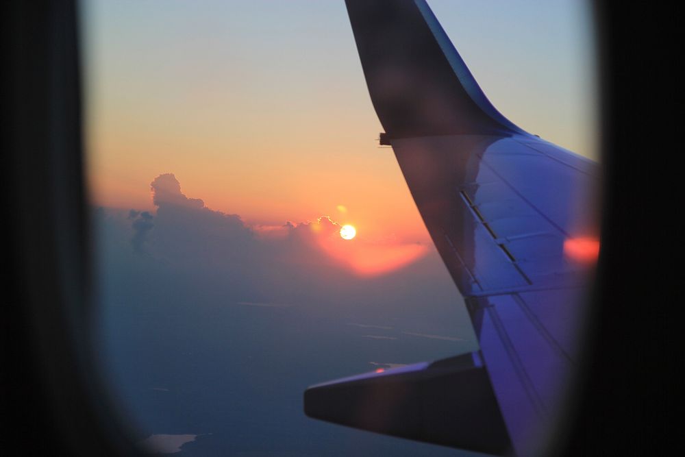 A passenger's view of the distance sunset from a plane window, with the plane's wing in the foreground. Original public…