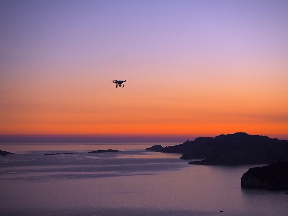A drone hovering over the water and rocks of La Ciotat with an orange and purple sunset in the background. Original public…