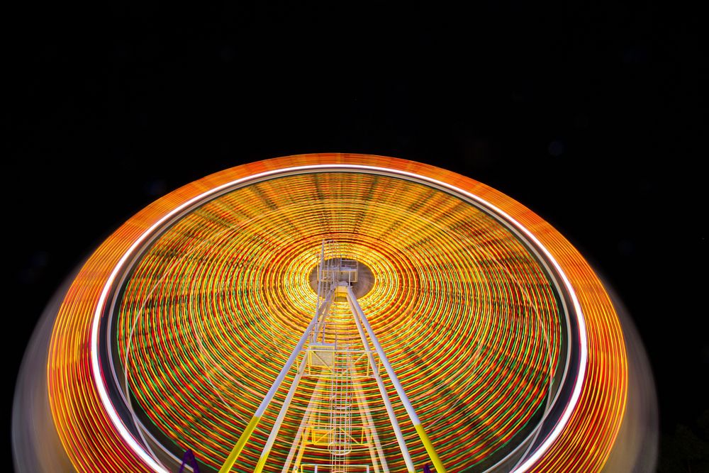 A time exposure shot of a ferris wheel with vivid orange and yellow light trails. Original public domain image from…