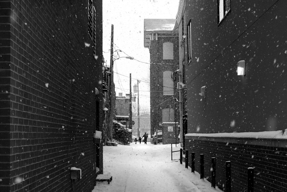 View from an alleyway in a city with snow falling around houses with a family walking through the street. Original public…
