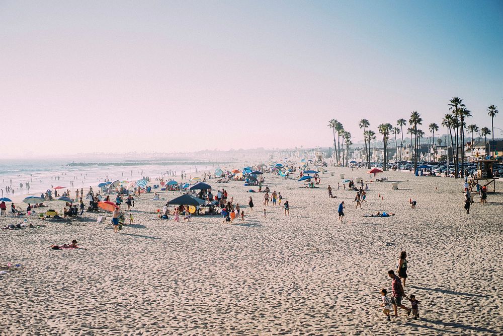 People enjoying the tropical sun, sea, and sand at a fairly crowded Newport Beach, CA. Original public domain image from…