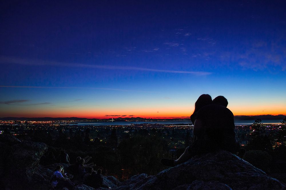 Couple in silhouette kisses atop a rocky hill, a city and sunset pictured in the background. Original public domain image…